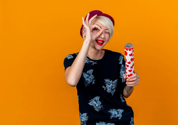 Impressionné jeune blonde party girl wearing party hat looking at camera holding confetti cannon faisant look geste isolé sur fond orange avec copie espace