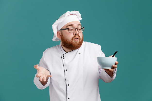 Impressionné jeune chef masculin portant des lunettes uniformes et une casquette tenant un bol vide avec une cuillère dedans montrant une main vide regardant un bol isolé sur fond bleu