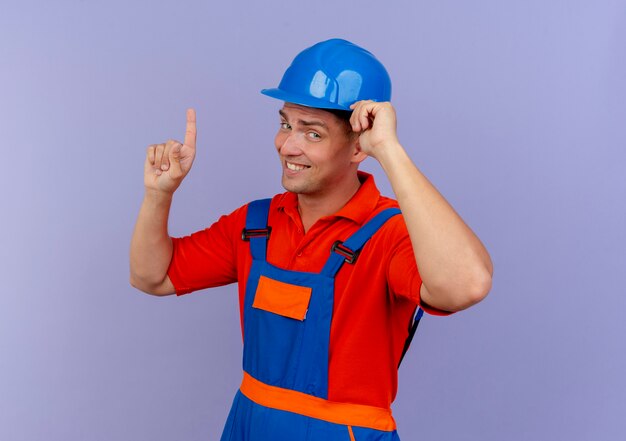 Impressionné jeune constructeur de sexe masculin portant l'uniforme et un casque de sécurité mettant la main sur le casque et pointe vers le haut