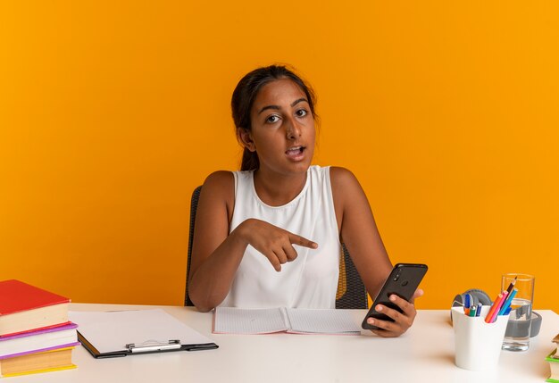 Impressionné jeune écolière assis au bureau avec des outils scolaires tenant et des points au téléphone