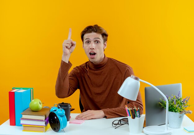 Impressionné jeune étudiant garçon assis au bureau avec des outils scolaires pointe vers le haut