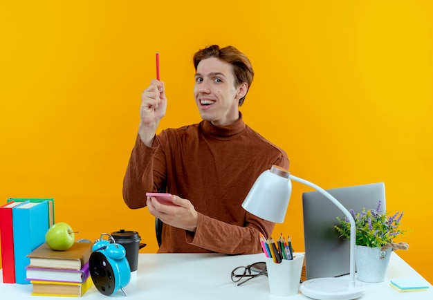 Impressionné jeune étudiant garçon assis au bureau avec des outils scolaires tenant un cahier et un stylo de levage