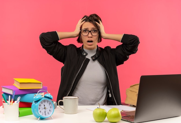 Photo gratuite impressionné jeune étudiante portant des lunettes assis au bureau à faire ses devoirs tenant sa tête isolée sur rose