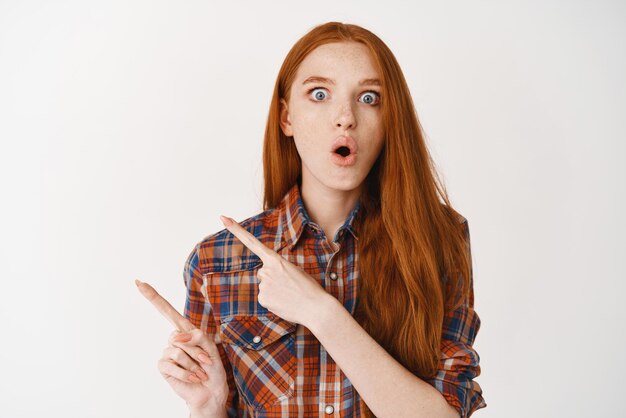 Impressionné jeune femme aux cheveux longs rouges et à la peau pâle regardant étonné et pointant du doigt à gauche la publicité debout sur fond blanc
