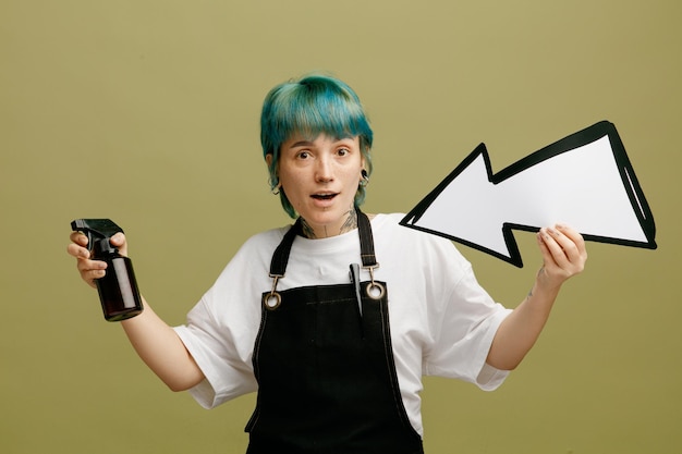 Impressionné jeune femme barbier portant l'uniforme tenant un fixatif à cheveux et une flèche pointant vers elle-même regardant la caméra isolée sur fond vert olive