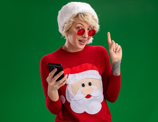 Photo gratuite impressionné jeune femme blonde portant chapeau de noël et pull de noël du père noël avec des lunettes tenant un téléphone mobile à la recherche et pointant vers le haut isolé sur un mur vert avec espace copie