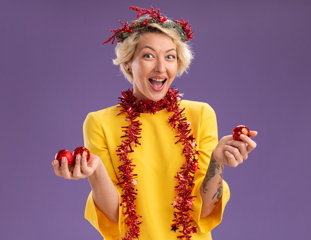 Impressionné jeune femme blonde portant une couronne de tête de Noël et guirlande de guirlandes autour du cou regardant la caméra tenant des boules de Noël isolé sur fond violet
