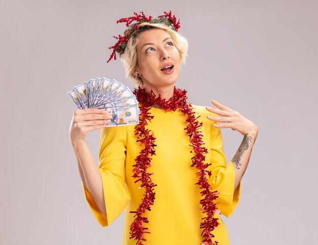 Impressionné jeune femme blonde portant une couronne de tête de Noël et guirlande de guirlandes autour du cou tenant de l'argent en gardant la main dans l'air en levant isolé sur mur blanc