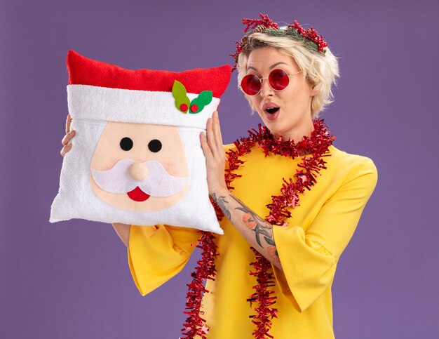 Impressionné jeune femme blonde portant une couronne de tête de Noël et guirlande de guirlandes autour du cou tenant l'oreiller du père Noël à isolé sur mur violet