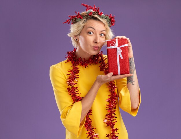 Impressionné jeune femme blonde portant une couronne de tête de Noël et guirlande de guirlandes autour du cou tenant un paquet cadeau à la recherche de lèvres pincées isolé sur mur violet avec espace copie