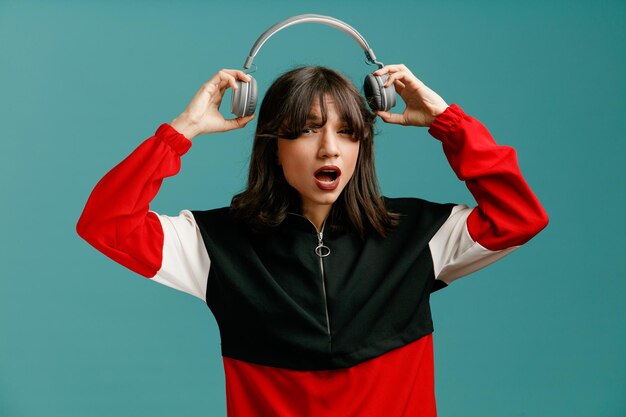 Impressionné jeune femme caucasienne regardant la caméra enlevant le casque isolé sur fond bleu