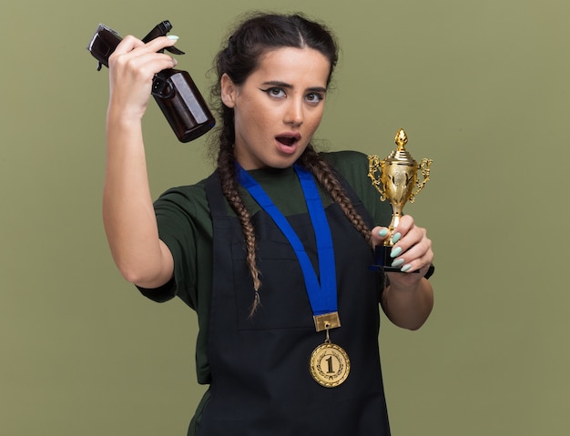 Impressionné jeune femme coiffeur en uniforme et médaille tenant la coupe du vainqueur et soulevant des outils de coiffeur isolés sur mur vert olive