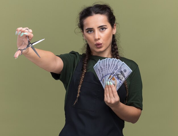 Impressionné jeune femme coiffeur en uniforme tenant de l'argent avec des ciseaux isolé sur mur vert olive