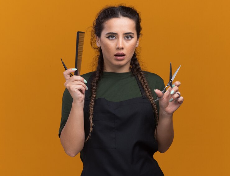 Photo gratuit impressionné jeune femme coiffeur en uniforme tenant des outils de coiffeur isolés sur un mur orange avec espace de copie