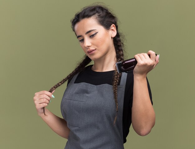 Impressionné jeune femme coiffeur en uniforme tenant une tondeuse à cheveux a attrapé les cheveux isolés sur mur vert olive