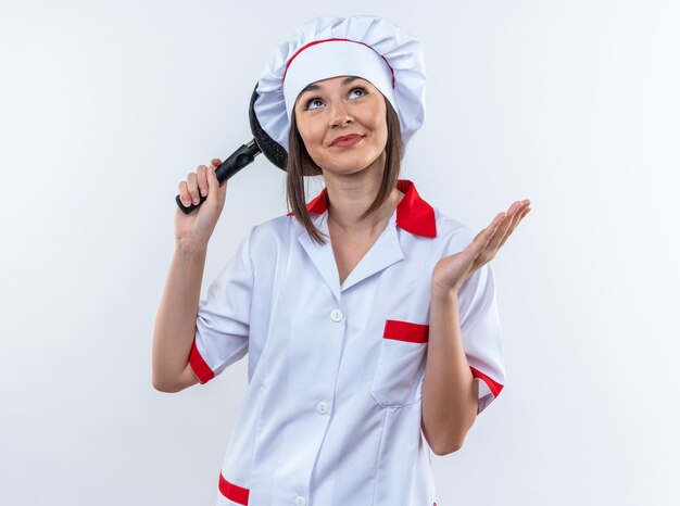 Impressionné jeune femme cuisinier portant l'uniforme de chef tenant une poêle à frire derrière la tête isolé sur fond blanc