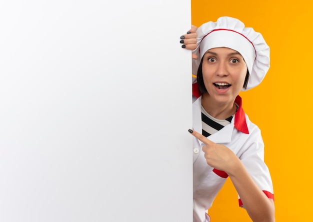 Photo gratuite impressionné jeune femme cuisinier en uniforme de chef à la recherche de derrière un mur blanc et pointant vers elle isolé sur orange avec copie espace