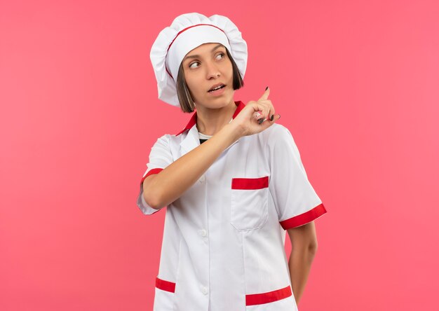 Impressionné jeune femme cuisinier en uniforme de chef regardant sur le côté et pointant derrière isolé sur rose avec copie espace