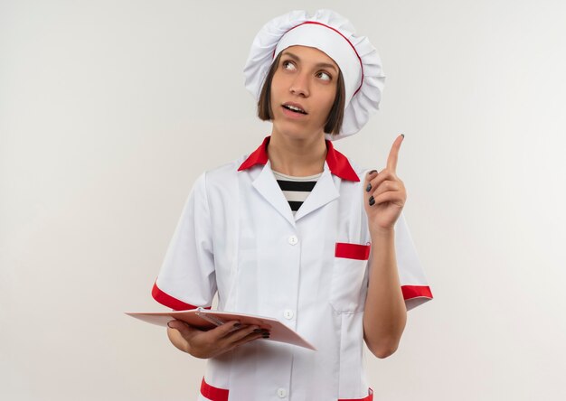 Impressionné jeune femme cuisinier en uniforme de chef tenant le bloc-notes et levant le doigt à côté isolé sur blanc avec espace copie