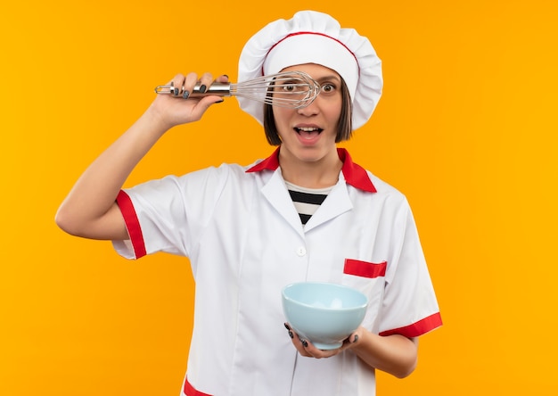 Impressionné jeune femme cuisinier en uniforme de chef tenant un fouet et un bol à travers le fouet isolé sur orange