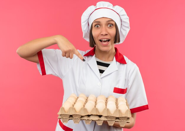 Impressionné jeune femme cuisinier en uniforme de chef tenant et pointant sur carton d'oeufs isolé sur rose