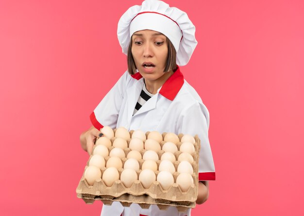 Impressionné jeune femme cuisinier en uniforme de chef tenant et regardant carton d'oeufs isolé sur rose avec copie espace