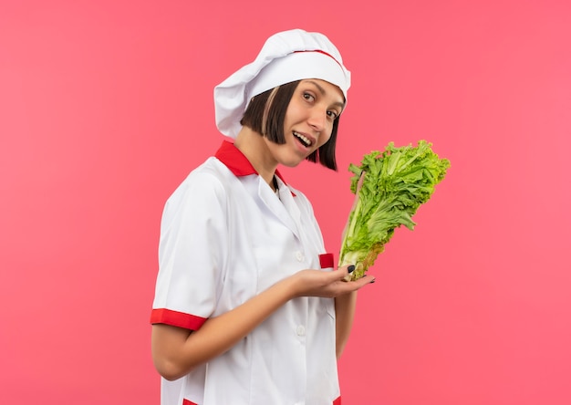 Impressionné jeune femme cuisinière en uniforme de chef tenant et pointant avec la laitue à la main isolé sur rose