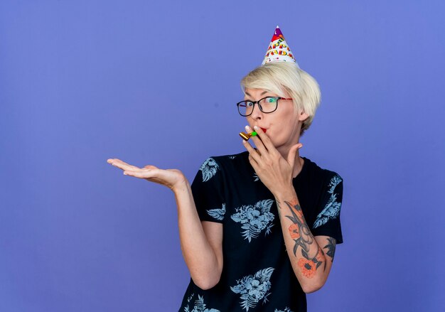 Impressionné jeune femme de fête blonde portant des lunettes et une casquette d'anniversaire à l'avant holding party blower dans la bouche montrant la main vide isolée sur le mur violet