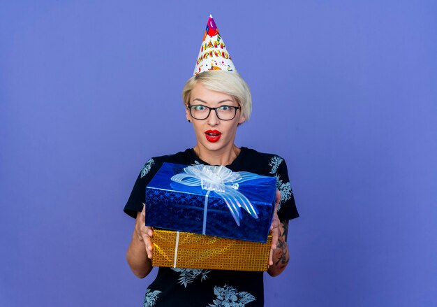 Impressionné jeune femme de fête blonde portant des lunettes et un chapeau d'anniversaire tenant des coffrets cadeaux à l'avant isolé sur mur violet