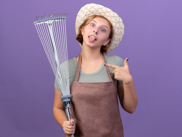 Impressionné jeune femme jardinier slave portant chapeau de jardinage sort la langue tenant et pointant sur le râteau à feuilles violet