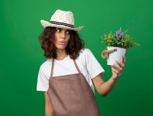 Impressionné jeune femme jardinière en uniforme portant chapeau de jardinage tenant et regardant la fleur en pot de fleurs