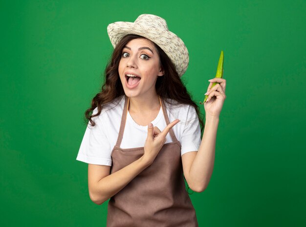 Impressionné jeune femme jardinière en uniforme portant chapeau de jardinage tient et pointe au piment isolé sur mur vert