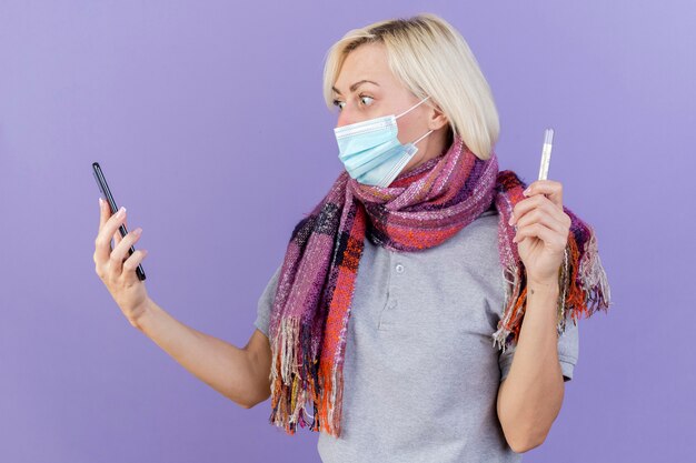 Impressionné jeune femme malade blonde portant un masque médical et une écharpe détient un thermomètre et se penche sur le téléphone isolé sur le mur violet