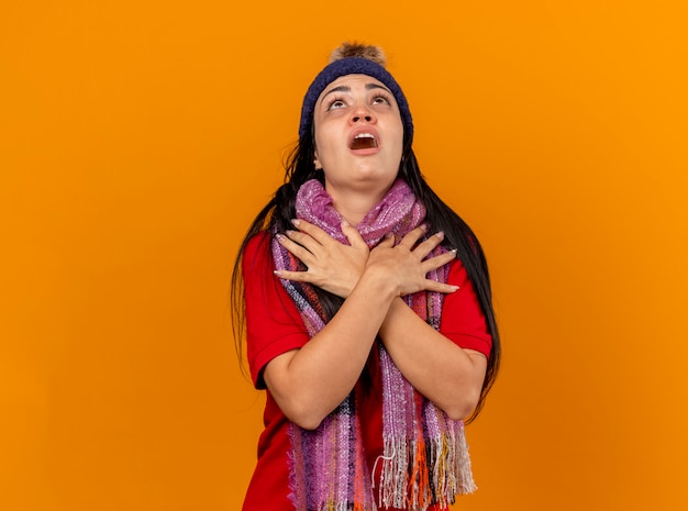 Impressionné jeune femme malade portant un chapeau d'hiver et une écharpe en gardant les mains croisées sur les épaules en levant isolé sur mur orange