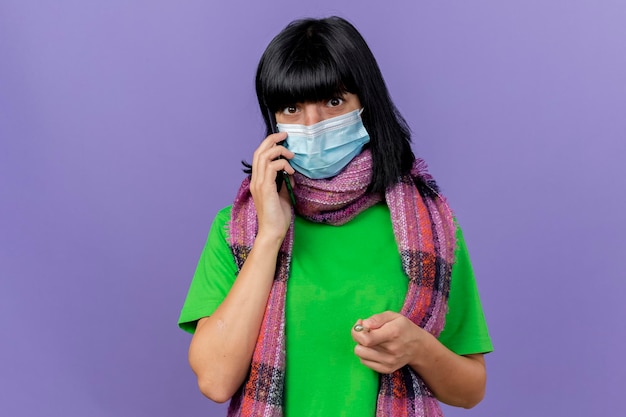 Photo gratuite impressionné jeune femme malade portant un masque et une écharpe à l'avant de parler au téléphone isolé sur un mur violet avec espace de copie