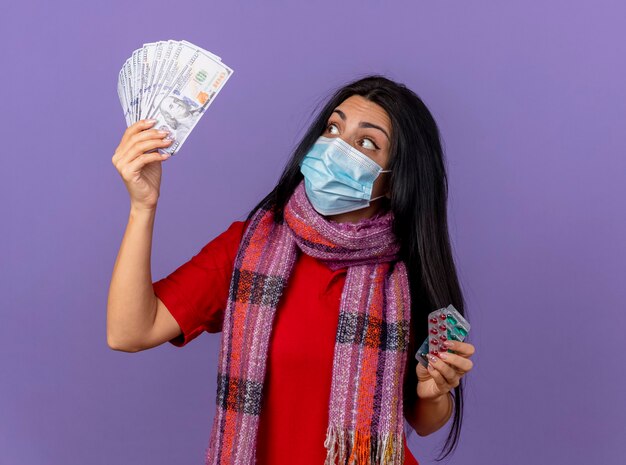 Impressionné jeune femme malade portant un masque et une écharpe tenant de l'argent et pack de capsules à l'argent isolé sur mur violet