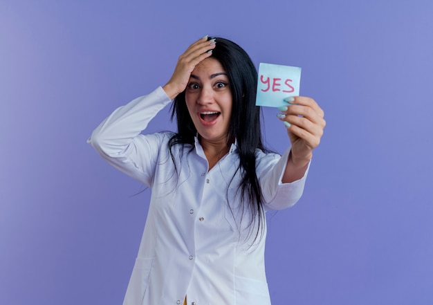 Impressionné jeune femme médecin portant une robe médicale montrant oui note, à garder la main sur la tête