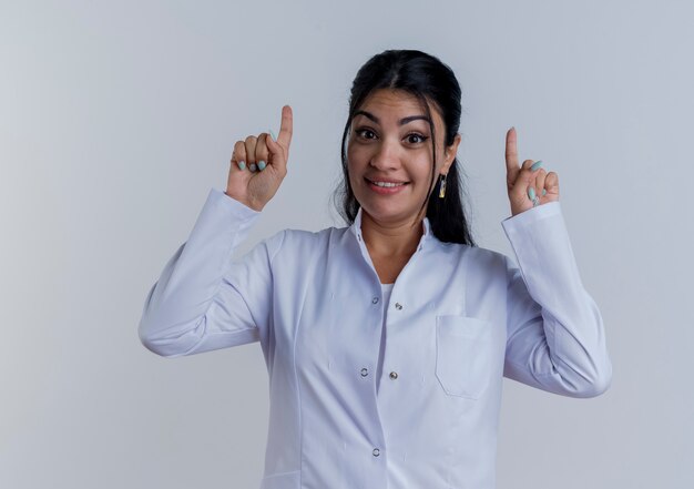 Impressionné jeune femme médecin portant une robe médicale pointant vers le haut isolé sur un mur blanc