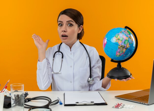 Impressionné jeune femme médecin portant une robe médicale et un stéthoscope assis au bureau avec des outils médicaux et un ordinateur portable tenant un globe et montrant une main vide