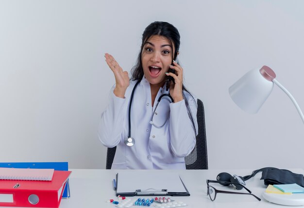 Impressionné jeune femme médecin portant robe médicale et stéthoscope assis au bureau avec des outils médicaux à la recherche de parler au téléphone montrant la main vide isolée