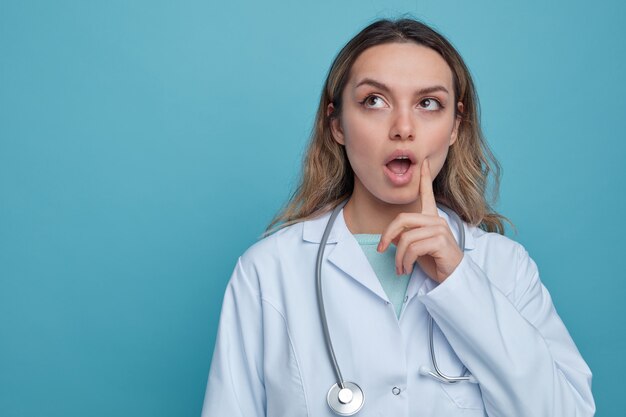 Impressionné jeune femme médecin portant une robe médicale et un stéthoscope autour du cou touchant la joue avec le doigt en levant