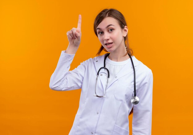 Impressionné jeune femme médecin portant une robe médicale et un stéthoscope en levant le doigt sur l'espace orange isolé avec copie espace