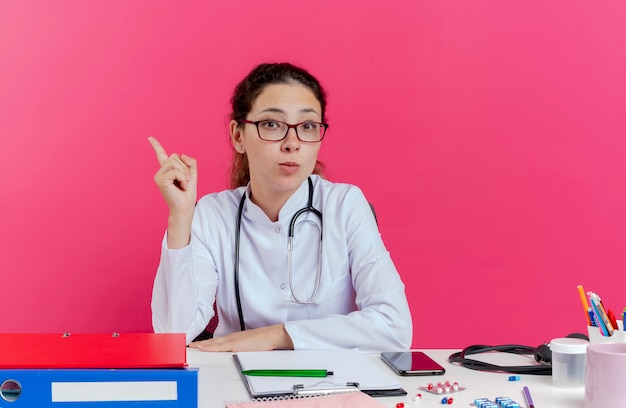 Impressionné jeune femme médecin portant une robe médicale et un stéthoscope et des lunettes assis au bureau avec des outils médicaux à la levée du doigt isolé