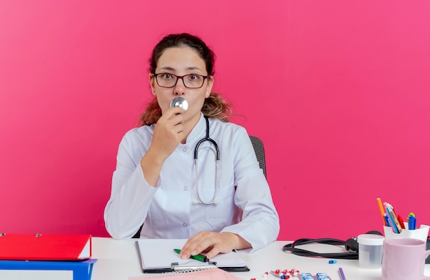 Impressionné Jeune Femme Médecin Portant Une Robe Médicale Et Un Stéthoscope Et Des Lunettes Assis Au Bureau