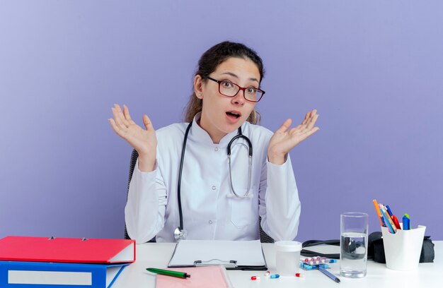 Impressionné jeune femme médecin portant une robe médicale et un stéthoscope à la recherche montrant les mains vides assis au bureau avec des outils médicaux isolés