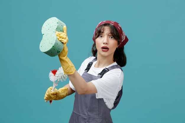 Impressionné jeune femme nettoyeuse portant un bandana uniforme et des gants en caoutchouc debout dans la vue de profil tenant des brosses de toilette étirant l'éponge regardant la caméra faire semblant de nettoyer