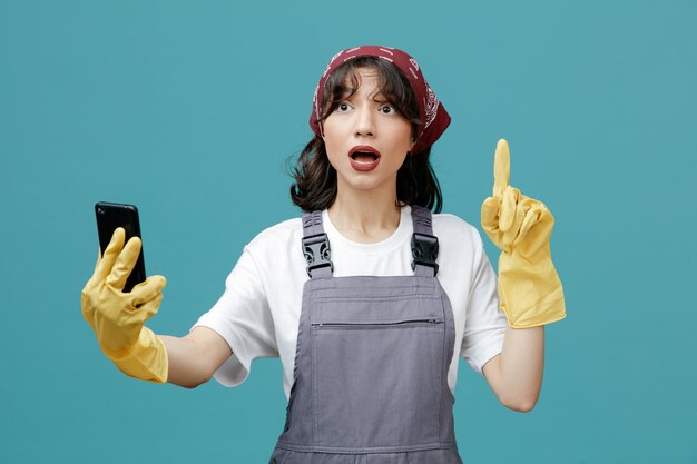 Impressionné jeune femme nettoyeuse portant un bandana uniforme et des gants en caoutchouc étirant le téléphone portable en regardant le côté pointant vers le haut isolé sur fond bleu