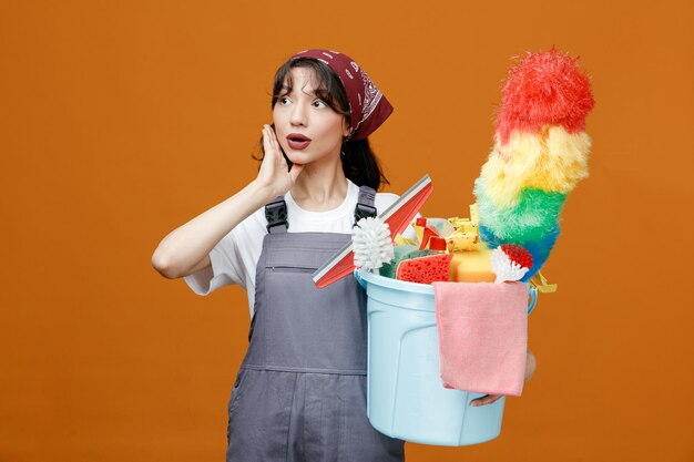 Impressionné jeune femme nettoyeuse portant l'uniforme et le bandana gardant la main sur le visage tenant un seau plein d'outils de nettoyage regardant le côté isolé sur fond orange