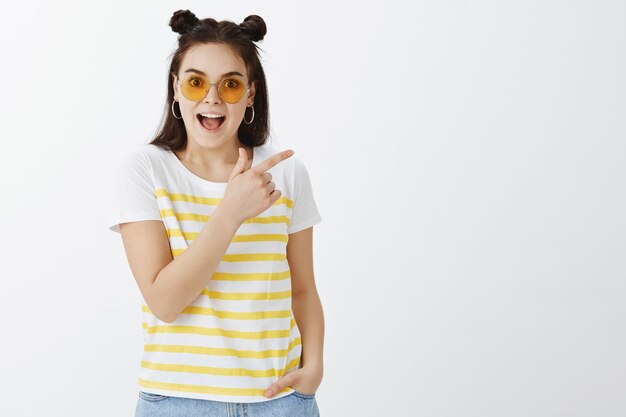 Impressionné jeune femme posant avec des lunettes de soleil contre le mur blanc