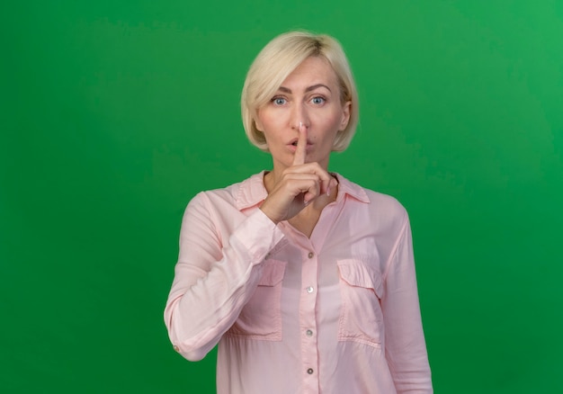 Impressionné jeune femme slave blonde faisant le geste de silence à la caméra isolée sur fond vert avec espace de copie